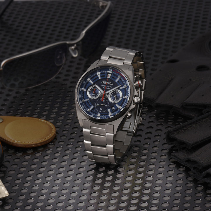 Het Seiko heren horloge SSB407P1, gemaakt van roestvrij staal, heeft een blauwe en zilveren wijzerplaat met subdials tegen een geperforeerd zwart oppervlak. Stijlvolle zonnebrillen, leren handschoenen en een beige leren sleutelhanger versterken dit strakke quartz uurwerk.