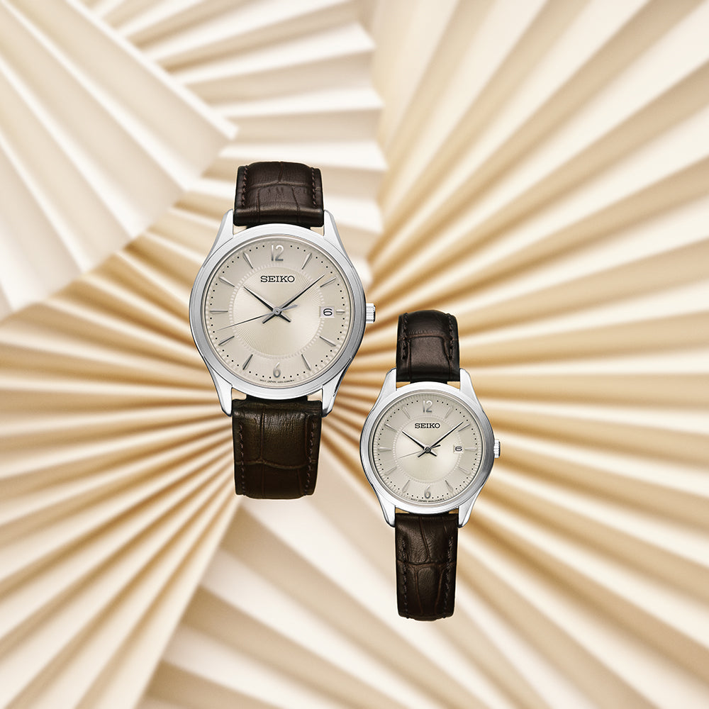 Twee Seiko SUR421P1 herenhorloges met bruine leren banden, zilverkleurige ronde kasten, eenvoudige beige wijzerplaten met uurmarkeringen, datumaanduiding, quartz uurwerk en saffierglas worden elegant tentoongesteld tegen een geometrische crèmekleurige achtergrond.