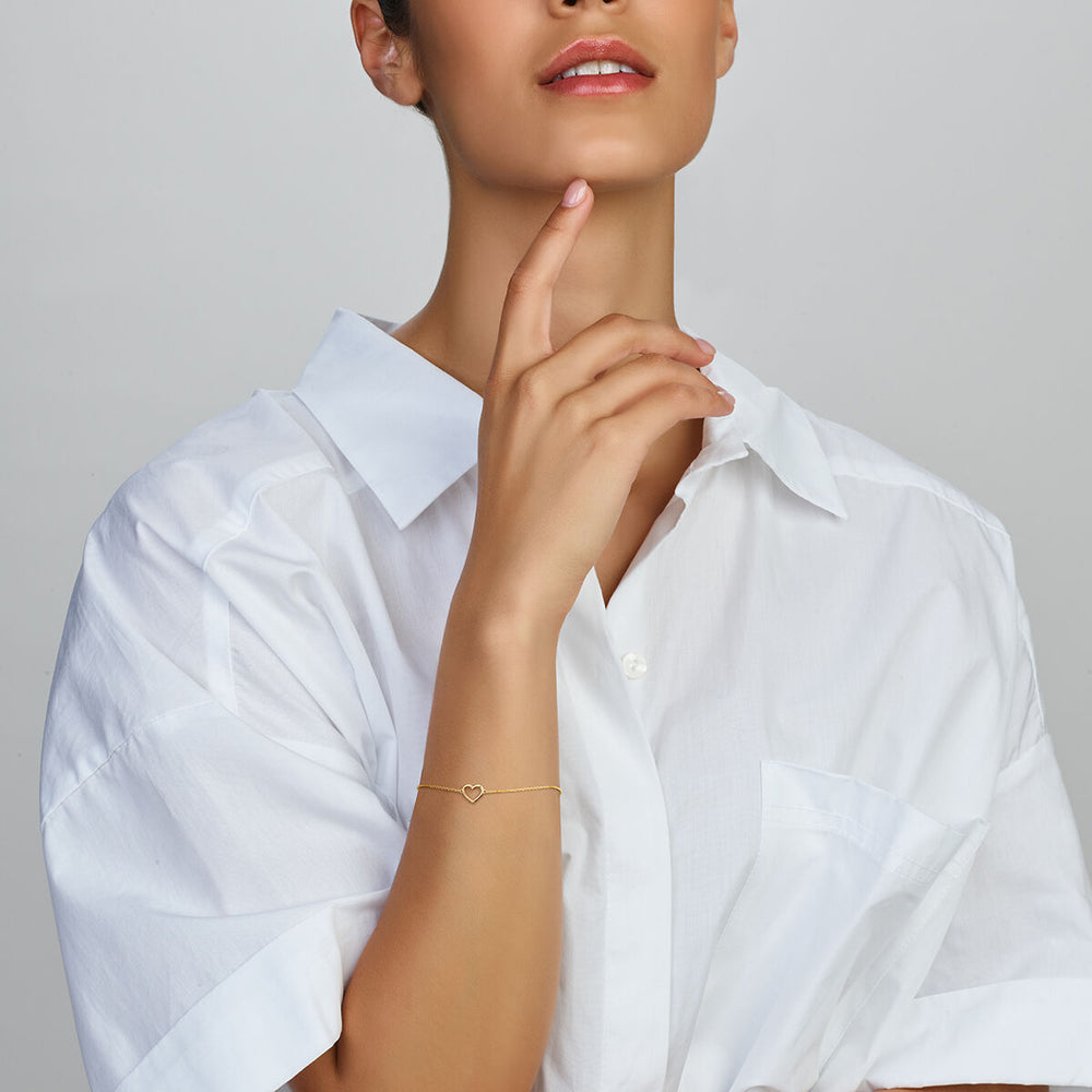 Een vrouw in een wit overhemd met kraag raakt zachtjes haar kin aan en toont elegant de Van der Kooij Collectie Gouden armband dames hart 14K om haar pols, waarbij deze delicate sieraad afsteekt tegen de neutrale achtergrond.