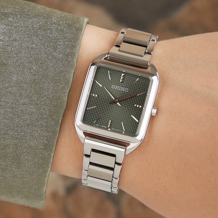 Een persoon draagt een Seiko dameshorloge SWR075P1 met een rechthoekige groene wijzerplaat, quartz uurwerk en metalen band. De wijzerplaat heeft zilveren uurmarkeringen en wijzers. De persoon is gekleed in een getextureerde groene mouw.