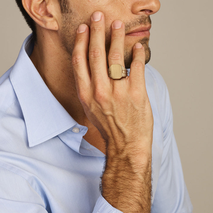Een man in een lichtblauw overhemd raakt zijn gezicht aan, terwijl hij hetveerring gematteerd 14K geelgoud van Van der Kooij Collectie laat zien. Zijn bedachtzame pose en de neutrale achtergrond benadrukken de elegante eenvoud van de ring.