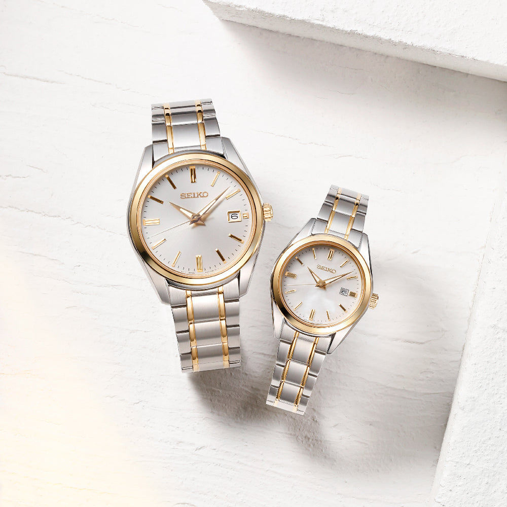 Twee Seiko dameshorloge SUR636P1 horloges met zilveren en gouden metalen banden en witte wijzerplaten worden gepresenteerd op een getextureerd wit oppervlak. Deze quartzhorloges hebben gouden uurmarkeringen, wijzers, kleine datumvensters en bieden 10ATM waterbestendigheid.