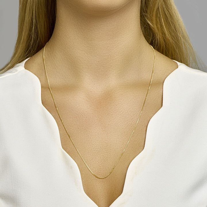 Een vrouw draagt een Van der Kooij Collectie collier gourmette 4-zijdes geslepen 1,2 mm 14K geelgoud met een witte geschulpte halslijn. De nadruk ligt op de ketting en de bovenborst, tegen een effen grijze achtergrond.