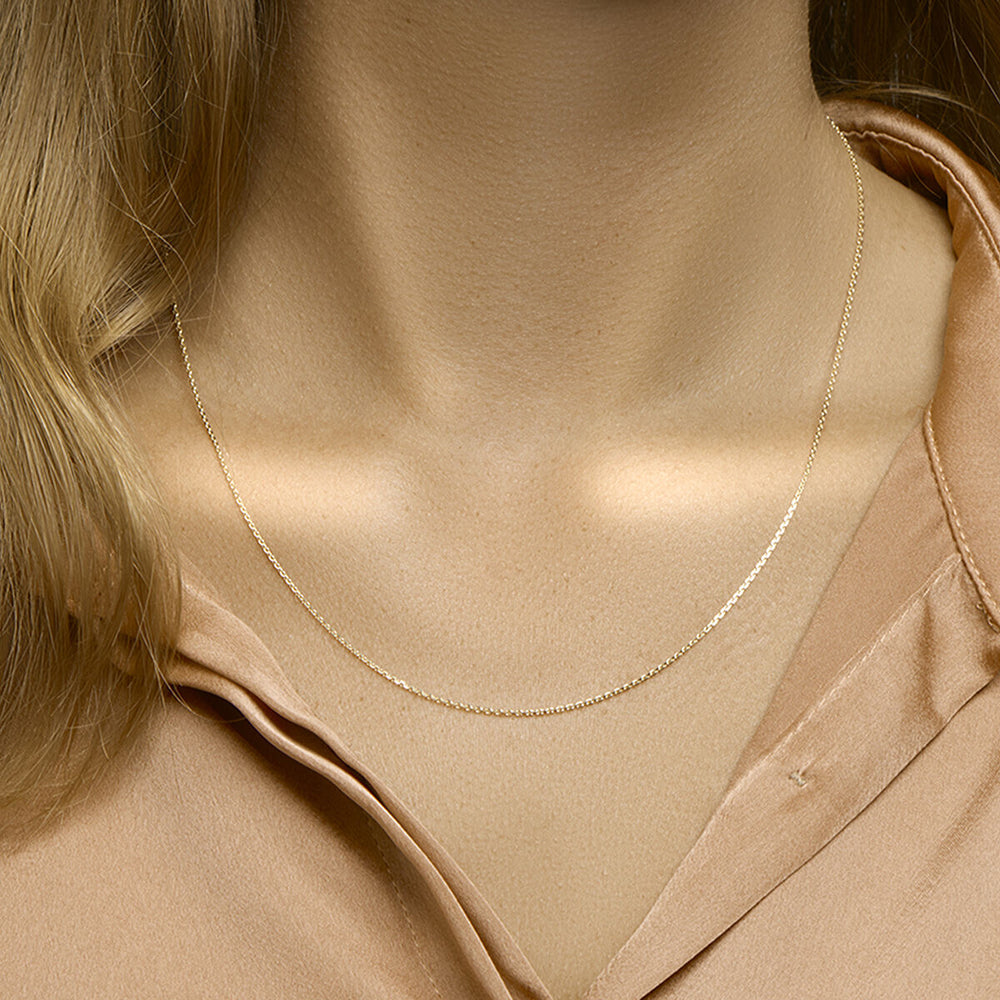Een close-up toont een persoon die de Van der Kooij Collectie collier anker gediamanteerd 1,1 mm 41 + 4 cm 3 micron zilver verguld (geel) ketting draagt over een lichtbruine satijnen blouse; alleen de hals, het sleutelbeen en een deel van de blouse zijn zichtbaar.
