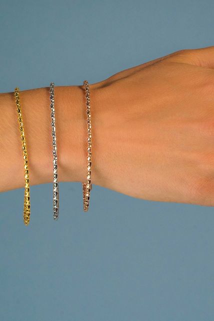 Een hand toont drie dunne, metaalachtige armbanden: goud, zilver en roségoud. De Zinzi Armband Zilver Zia1129 A heeft een delicaat kettingontwerp tegen een effen blauwe achtergrond, wat elegantie toevoegt aan elke outfit.