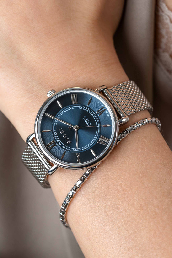 Close-up van een pols met het Zinzi zilverkleurige Horloge Ziw2053 met een zilveren meshband en blauwe wijzerplaat met Romeinse cijfers, gecombineerd met een dunne zilveren gevlochten band.