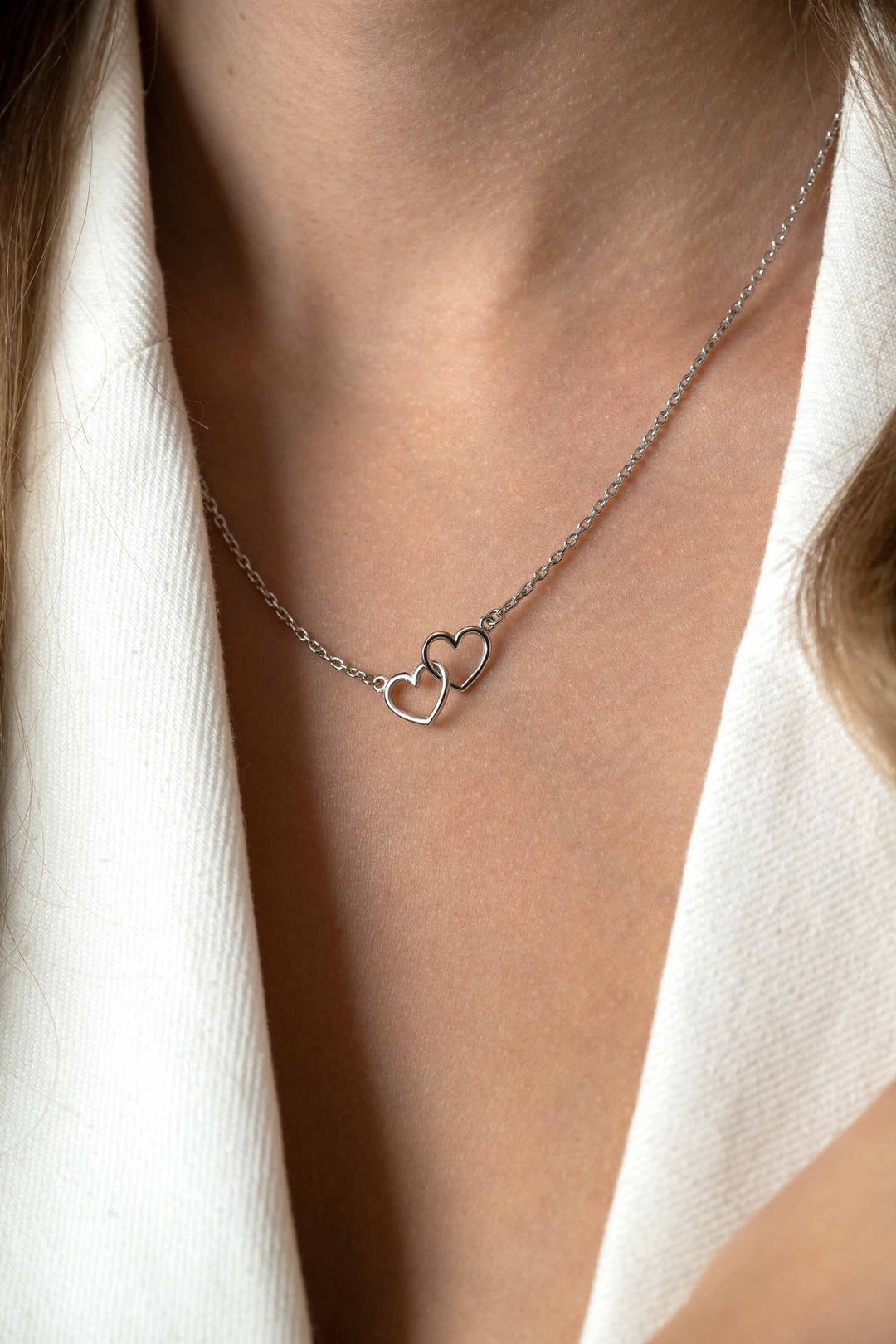 Een close-up van een persoon die de Zinzi Ketting Zilver Zic2513 van Zinzi draagt, met twee in elkaar grijpende hartvormige hangers, afgebeeld tegen een lichtgekleurde top met V-hals.
