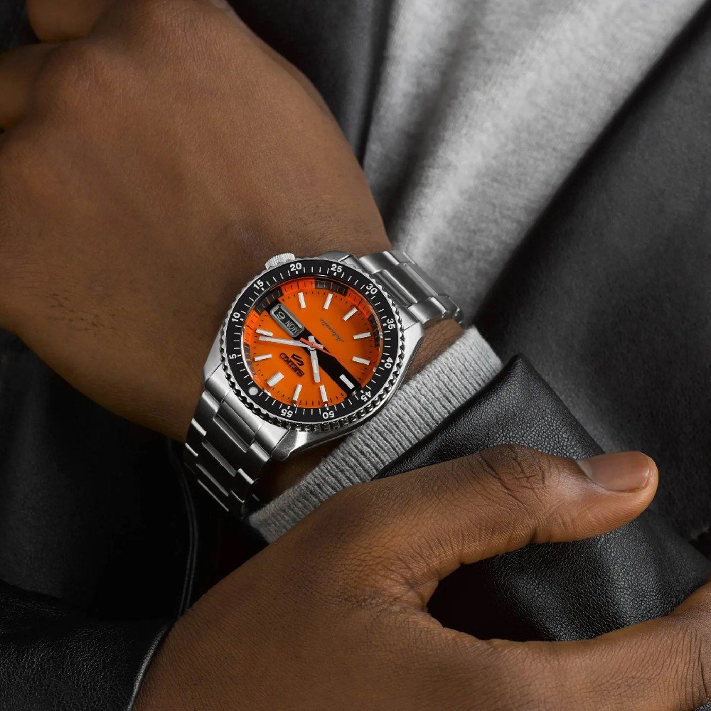 Een persoon controleert nonchalant de tijd op zijn Seiko 5 Sports SRPK11K1, een automatisch horloge met een feloranje wijzerplaat en dag/datumweergave, met een waterdichtheid van 10ATM. Ze zijn gekleed in een grijs shirt en een zwart leren jack.