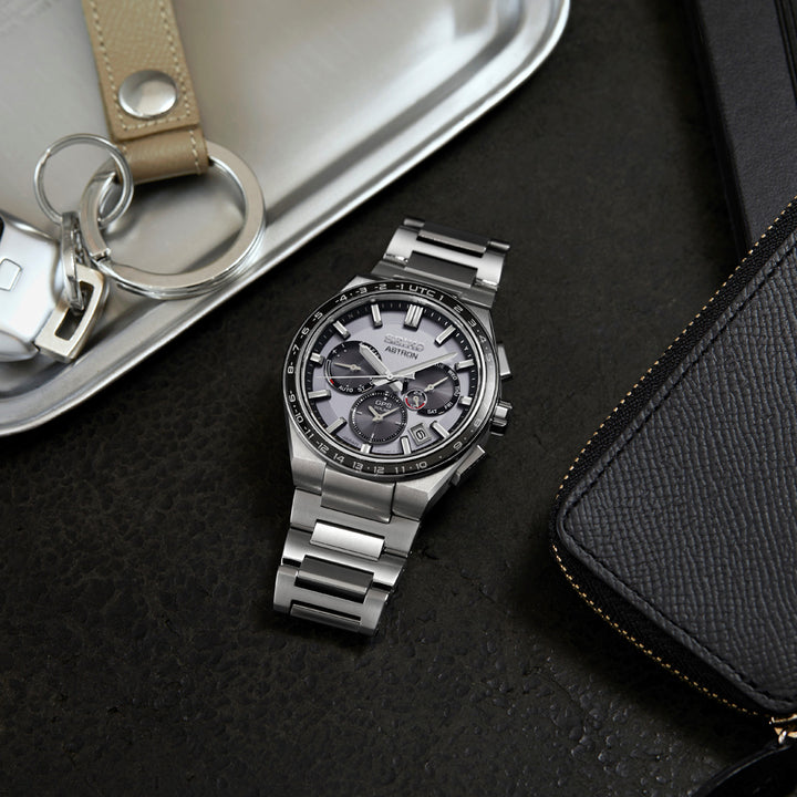 Een zilveren Seiko Astron SSH109J1 herenhorloge met een metalen band rust op een donker oppervlak naast een zwarte leren portemonnee. Een metalen lade op de achtergrond bevat sleutels met een beige sleutelhanger. Het duurzame saffierglas van het horloge voegt een elegante touch toe.