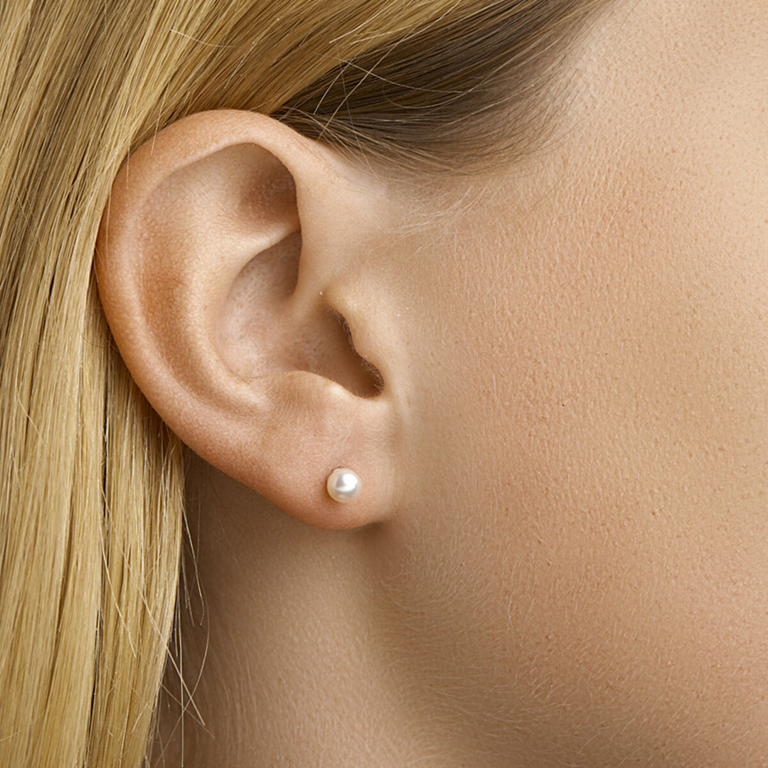 Close-up van het oor van een vrouw met steil blond haar, gekleed in Van der Kooij Collectie oorknoppen van parelmoer in 14 karaat geelgoud. De gladde huid accentueert de kleine, ronde witte parel in het midden van haar oorlel.