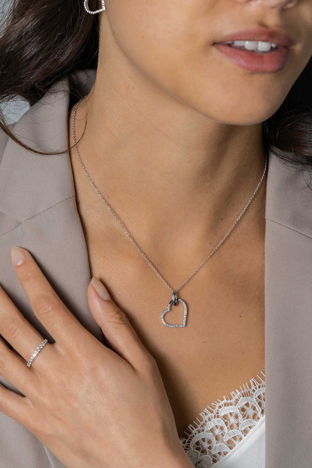 Een vrouw in een beige blazer en wit kanten topje showt de Zinzi Hangers Zilver ZIH2447 in een luxe hartontwerp, gecombineerd met een bijpassende zilveren ring versierd met witte zirkonia's, haar hand zachtjes rustend op haar borst.