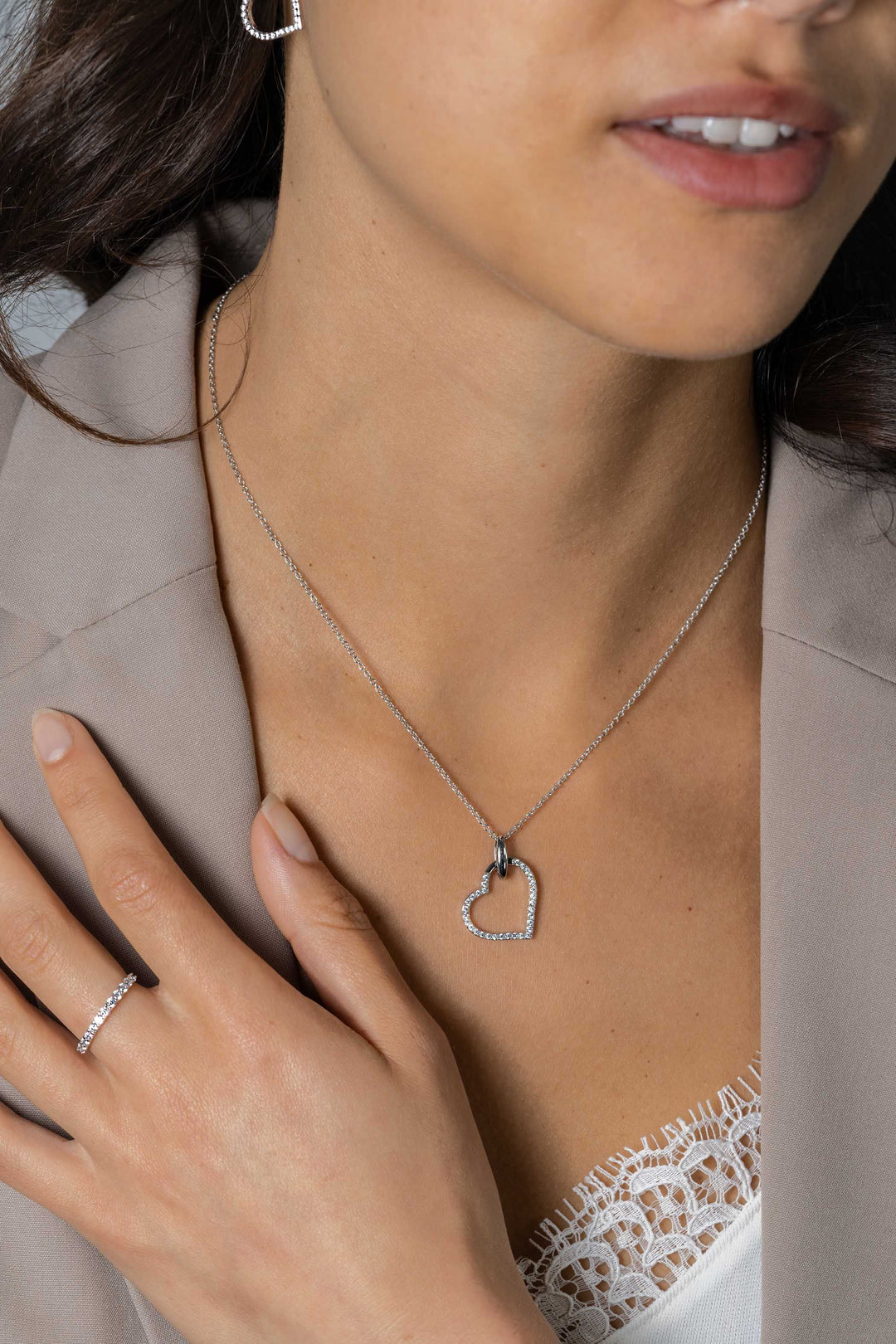 Een vrouw in een beige blazer en wit kanten topje showt de Zinzi Hangers Zilver ZIH2447 in een luxe hartontwerp, gecombineerd met een bijpassende zilveren ring versierd met witte zirkonia's, haar hand zachtjes rustend op haar borst.