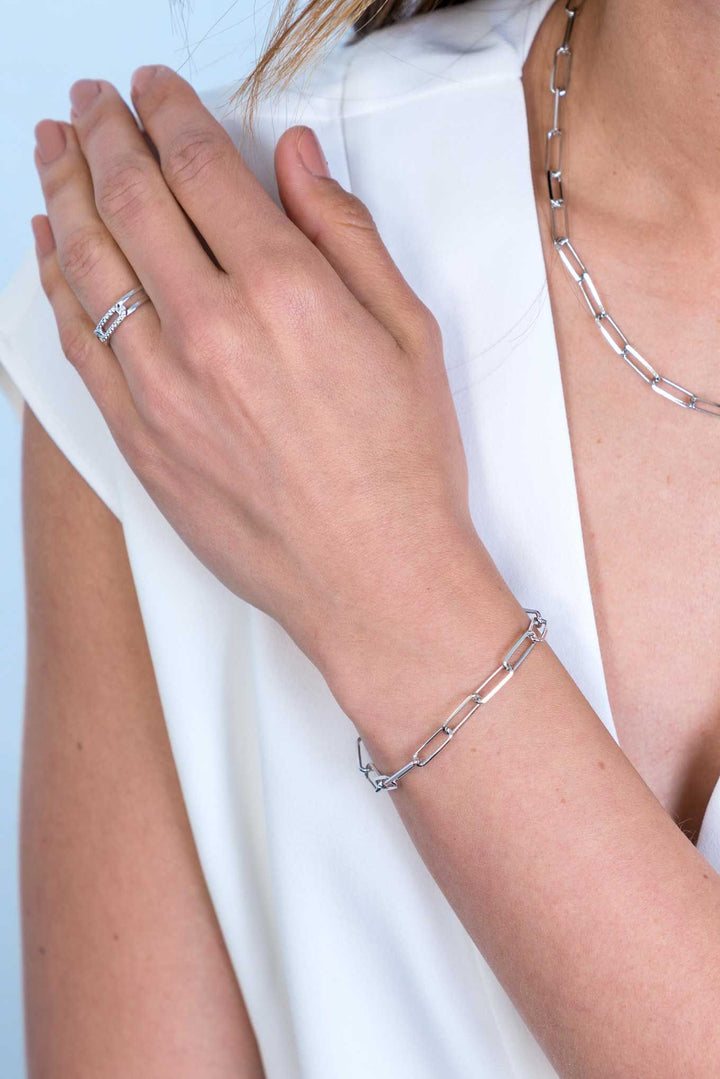 Een persoon die een witte blouse draagt, benadrukt verbluffende Zinzi-sieraden, waaronder de zilveren Zinzi Armband Zilver Zia2207, met gekoppelde rechthoekige kettingontwerpen als een paperclip. De elegante ketting, armband en ring vallen op tegen de lichte achtergrond.