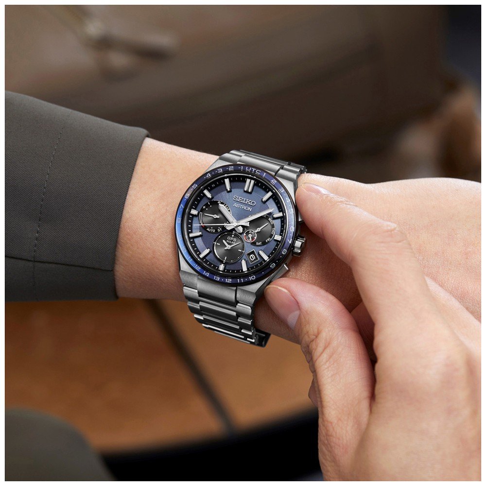 Een persoon stelt de tijd in op een Seiko Astron heren horloge SSH109J1. Dit zilver en blauwe horloge, onderdeel van de Astron-collectie, omvat drie sub-wijzerplaten, een datumaanduiding, lichtgevende wijzers onder saffierglas, en dit alles terwijl het een donkergroene mouw draagt.