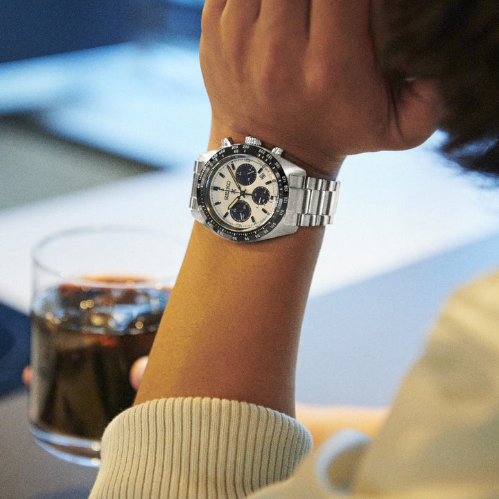 Een persoon die een Seiko Prospex heren horloge SSC813P1 draagt, met zijn opvallende wit-zwarte wijzerplaat, rust zijn hand bij een glas donkere vloeistof. Het elegante Saffierglas-ontwerp valt op en vestigt de aandacht op het solar uurwerk horloge en de zacht wazige achtergrond.