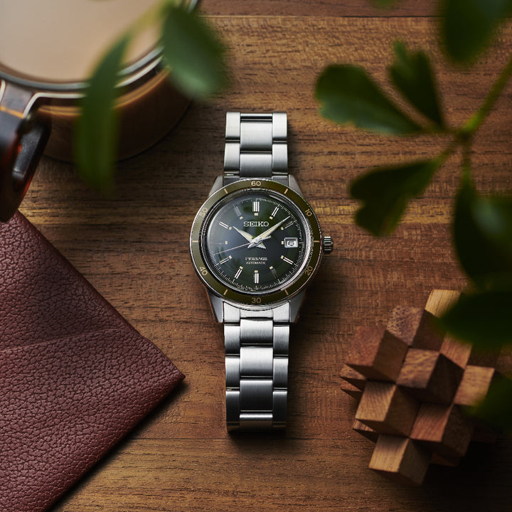 Het Seiko Presage herenhorloge SRPG07J1, met een groene wijzerplaat en datumaanduiding, staat op hout. In de buurt bevinden zich een leren portemonneehoek, een houten puzzel, een dekselbeker en groene plantenbladeren. Dit roestvrijstalen horloge uit de Presage-collectie van Seiko beschikt over een automatisch uurwerk.