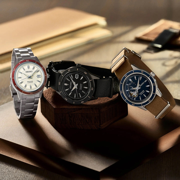 Drie polshorloges rusten op hout: Seiko met een witte wijzerplaat en zilveren band, een strak automatisch opwindmodel met Hardlex-glas en een horloge met een blauwe wijzerplaat en een bruine band. Notitieboekjes en een pen sieren elegant de achtergrond.