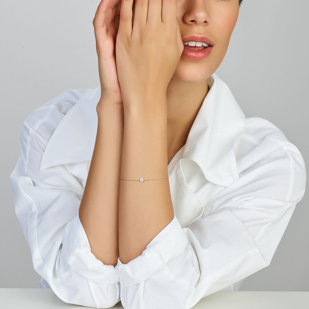 Een vrouw in een wit shirt poseert, haar gezicht deels bedekkend, terwijl ze de Van der Kooij Collectie Gouden armband dames zirkonia 14K met een subtiele ronde zirkonia bedel om haar pols draagt.