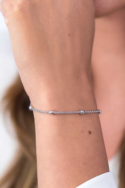 Een close-up toont een pols die de Zinzi Armband Zilver Zia1128 draagt, met een delicate zilveren ketting met kleine kralen. Een moedervlek op de huid voegt charme toe tegen een zacht wazige achtergrond.