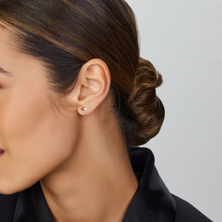 In profiel is een vrouw met donker haar in een lage knot afgebeeld, gekleed in Van der Kooij Collectie oorknoppen bolletje zilver gerhodineerd en een zwart shirt, waarbij de eenvoudige zilveren studs tegen een effen, lichte achtergrond zijn geplaatst.