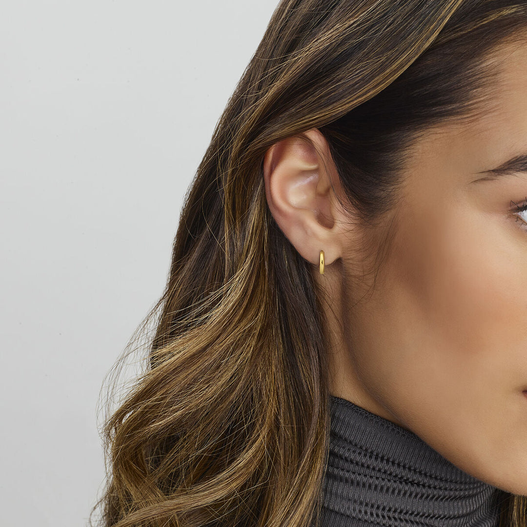 Close-up van het rechteroor van een vrouw met Van der Kooij Collectie klapoorringen 14K geelgoud met scharniersluiting. Ze heeft lang, golvend bruin haar, een grijze geribbelde coltrui en de achtergrond is effen en lichtgekleurd.