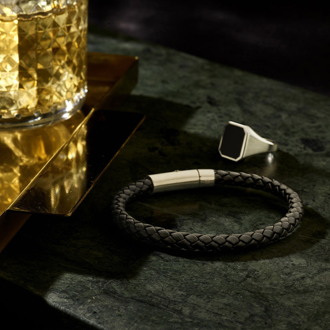 Een gevlochten zwart leren armband en een Van der Kooij Collectie zegelring massief onyx 13 x 11mm zilver gerhodineerd rusten op donkergroen marmer naast een glas whisky op een gouden dienblad.