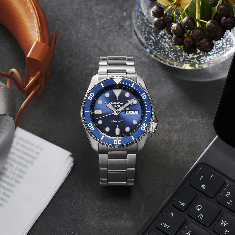 Naast een laptop, koptelefoon, papieren en een schaal met bessen staat het Seiko 5 Sports heren horloge SRPD51K1. Dit zilveren horloge van Seiko heeft een blauwe wijzerplaat die elegant de dag en datum weergeeft.