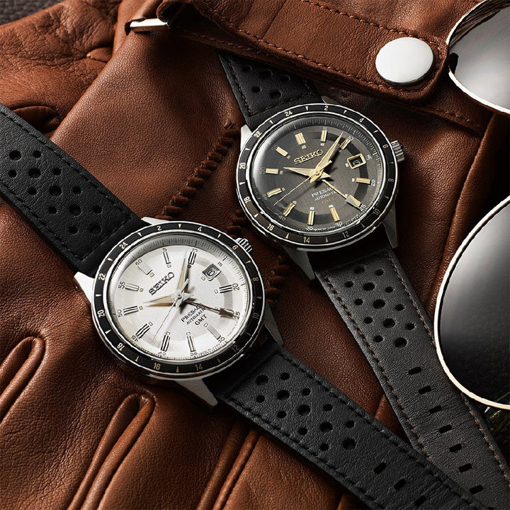 Twee Seiko Presage heren horloges, met zwarte leren bandjes, rusten op een bruine leren handschoen. De ene heeft een witte wijzerplaat, de andere een zwarte. Zonnebrillen met metalen montuur voegen subtiele elegantie toe.