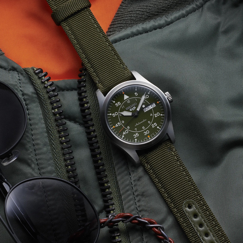 Een groen Seiko 5 Sports herenhorloge SRPH29K1 in militaire stijl met een nylon band wordt getoond op een oranje gevoerde jas met een gedeeltelijk open rits. Zwarte zonnebrillen met een gevlochten band complementeren de opstelling en versterken de robuuste uitstraling van dit horloge-ensemble van Seiko.