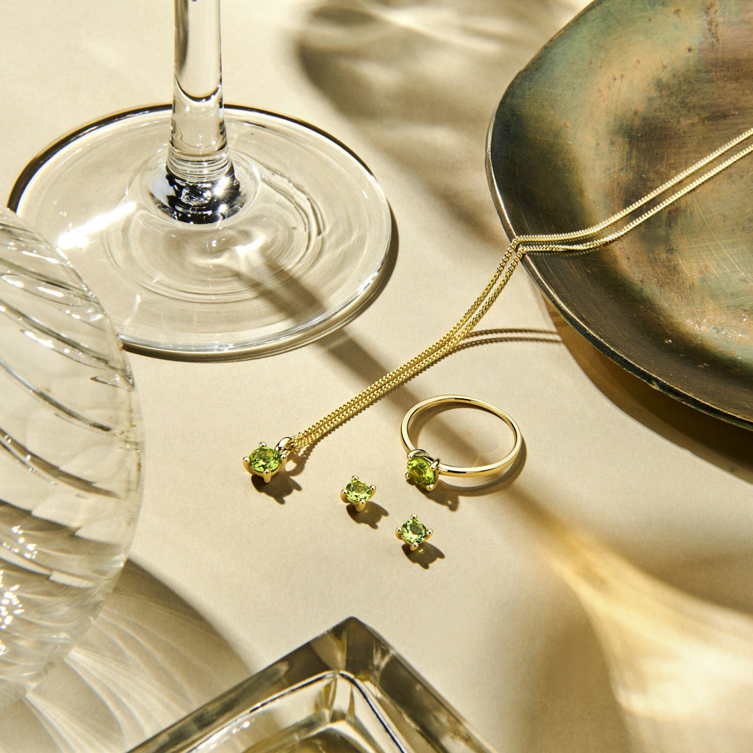 Gouden collier, ring en oorbellen met groene edelstenen, waaronder de Van der Kooij Collectie hanger peridot 14K geelgoud, gepresenteerd op beige naast een wijnglas, structuurglas en metalen schaal. Zonlicht creëert zachte schaduwen en reflecties.