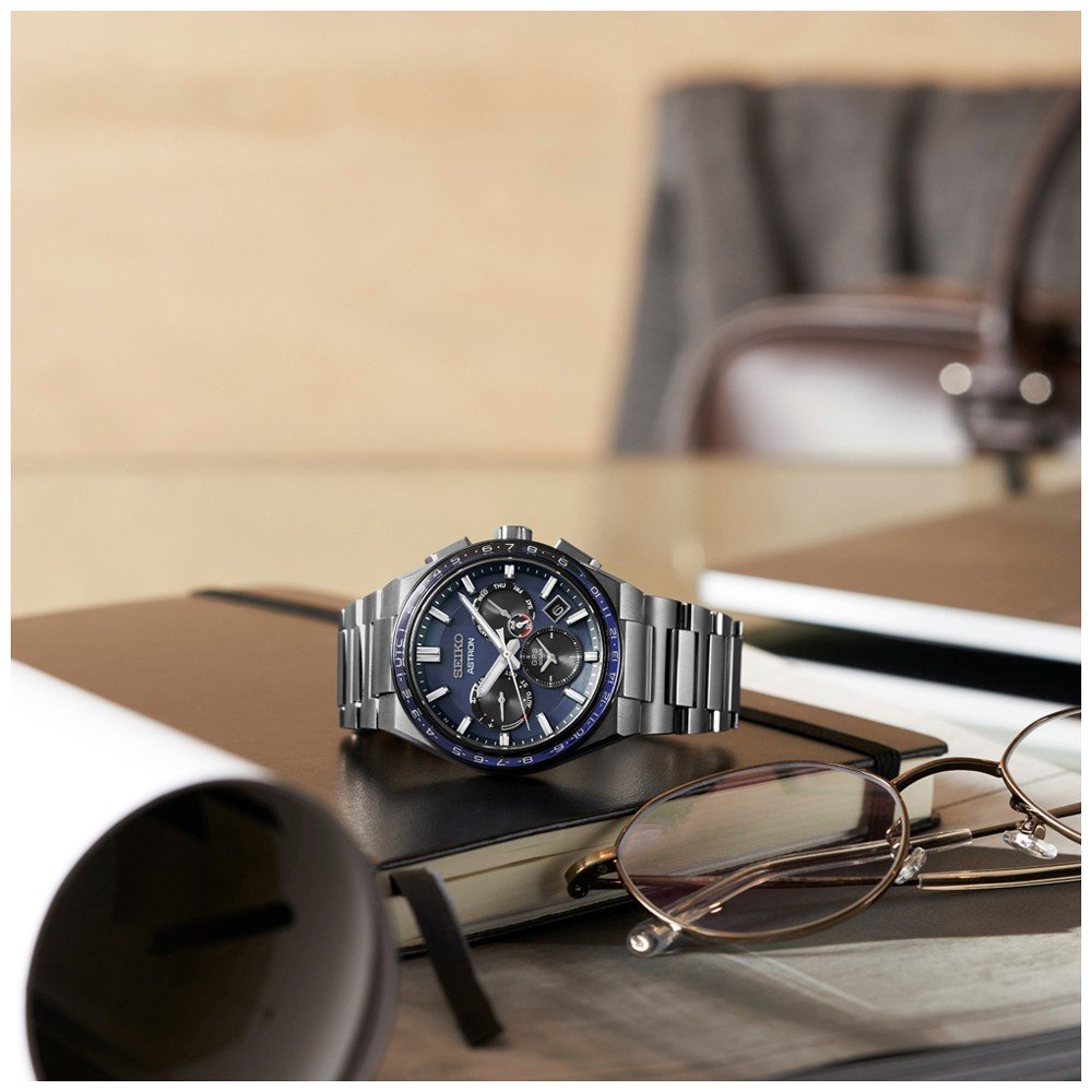 Het Seiko Astron heren horloge SSH109J1 met een blauwe wijzerplaat en saffierglas rust op zwarte notitieboeken op een glazen tafel, vergezeld door een bril en een opgerold document. Een stoel op de achtergrond maakt de professionele kantoorscène compleet.