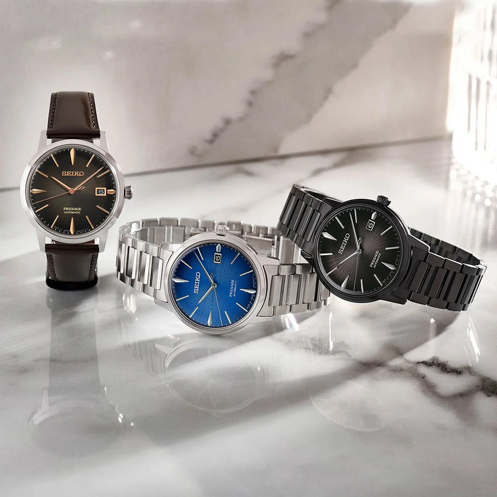 Drie Seiko horloges staan op een marmeren oppervlak: een met een bruine leren band en zwarte wijzerplaat, de Seiko Presage SRPJ13J1 met een zilveren metalen band, roestvrijstalen kast, blauwe wijzerplaat en een grijs metalen band automatisch horloge in het zwart. Zonlicht schaduwt zachtjes over de scène.