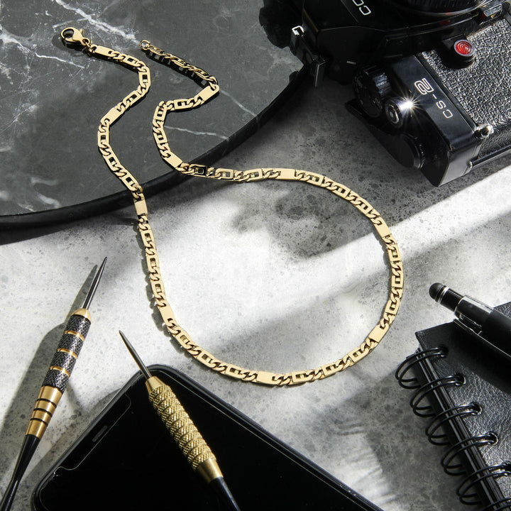 De Gouden ketting heren - collier valkenoog 4,7 mm 14K van Van der Kooij Collectie ligt op marmer naast een camera, potlood, pen, notitieboek en smartphone; licht en schaduw geven de scène robuuste elegantie.
