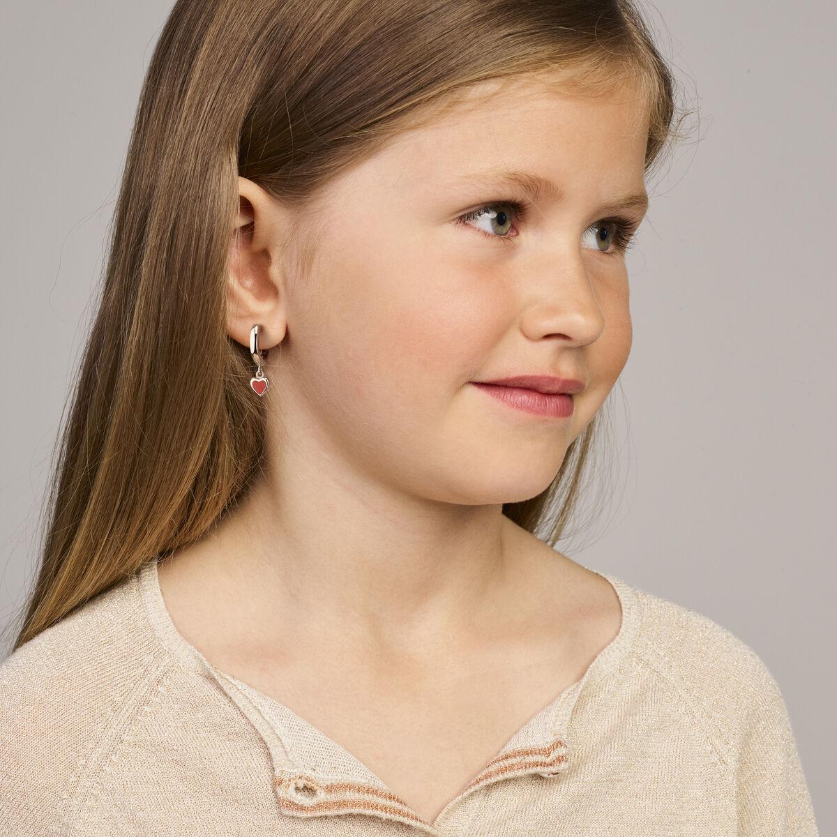 Een jong meisje met lang lichtbruin haar in een beige topje glimlacht zachtjes naar haar toe. Ze draagt een Van der Kooij Collectie klapoorringen hart rood emaille 18 mm zilver gerhodineerd. De achtergrond is effen en lichtgrijs.