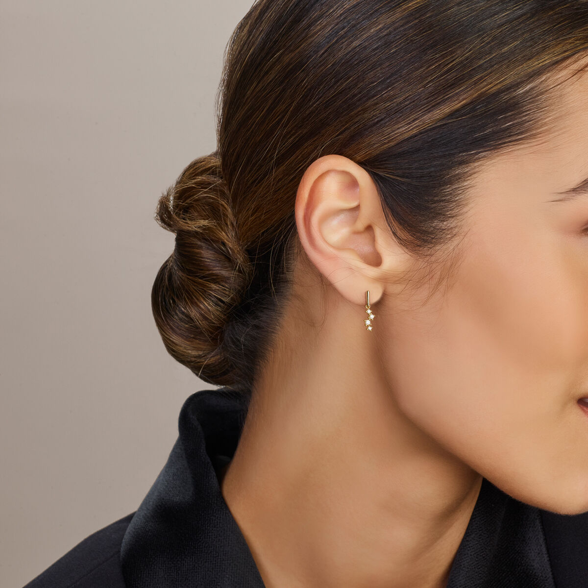 Een vrouw met donker haar in een nette lage knot draagt Van der Kooij Collectie oorhangers met zirkonia in 14 karaat geelgoud. Gekleed in een zwarte top wordt ze van opzij getoond tegen een neutrale achtergrond, waardoor deze elegante damessieraden extra goed tot hun recht komen.