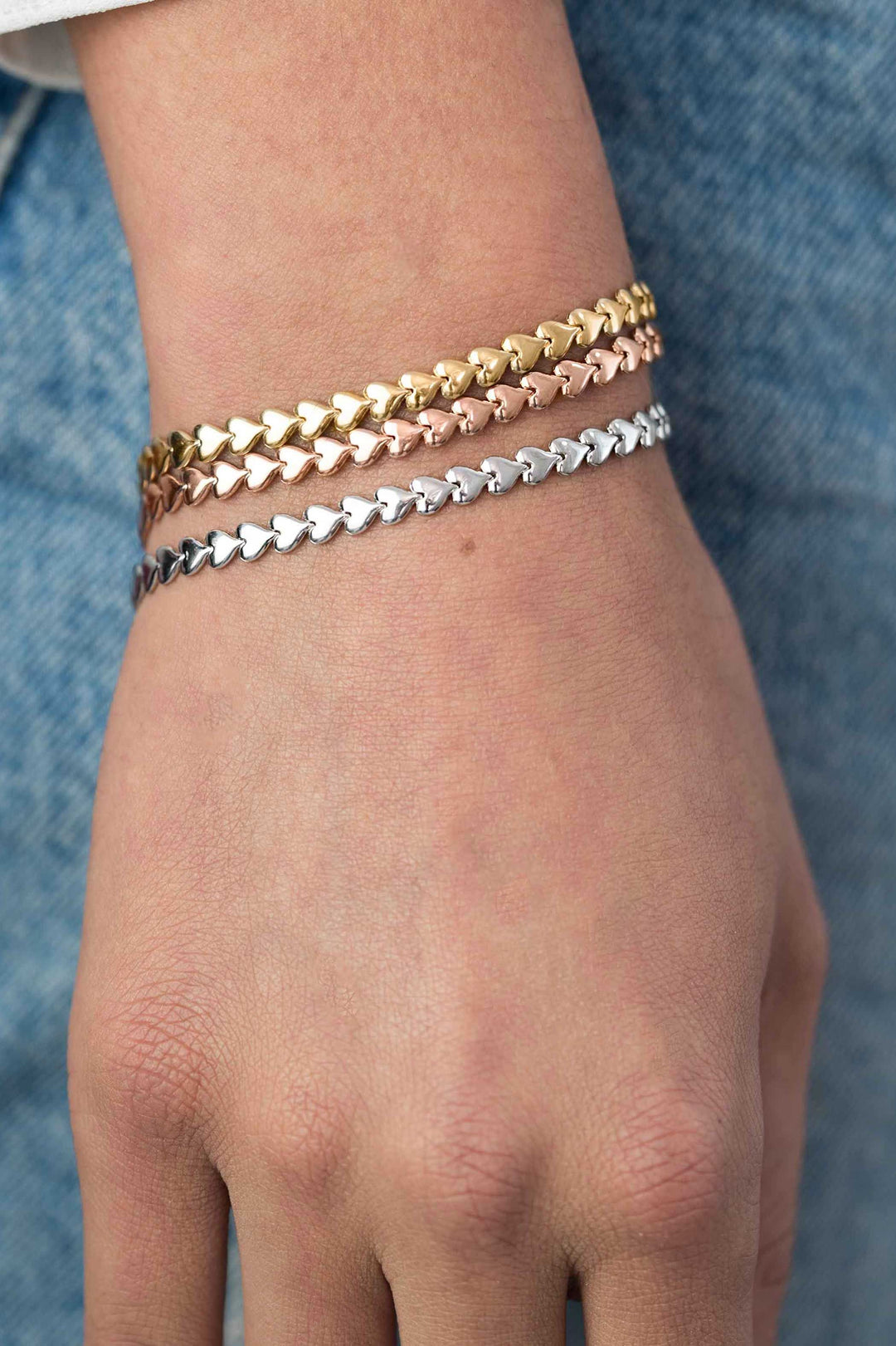 Een close-up van een hand die drie hartvormige kettingarmbanden draagt, waaronder de Zinzi Armband Zilver Zia1798 van Zinzi in goud-, roségoud- en zilvertinten, tegen een blauwe spijkerbroek.