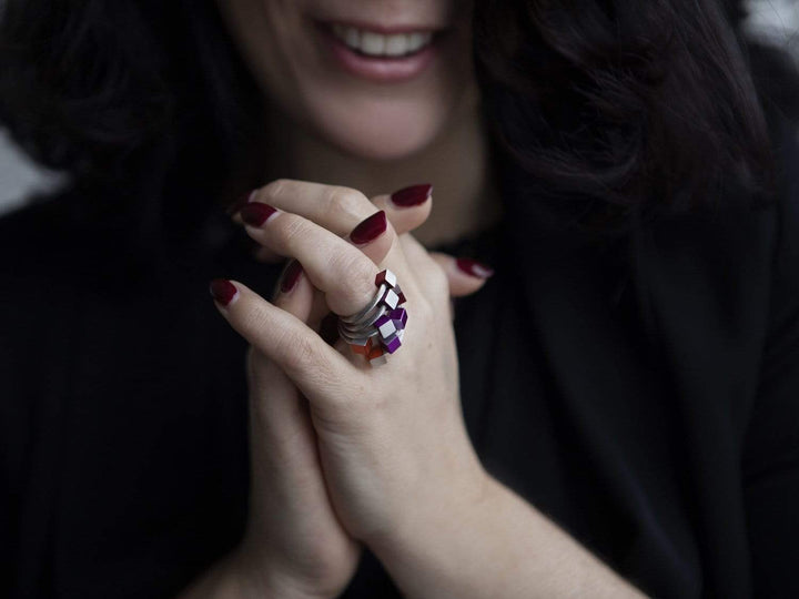 Een persoon met donker haar en bordeauxrode nagellak vouwt zijn handen en toont CLIC Creations Ring Kubusjes Rood R4R. Gemaakt van chirurgisch edelstaal in paarse en zilveren geometrische vormen, benadrukken de handgemaakt in Nederland ringen een zachte glimlach en complementeren ze perfect een donkere outfit.