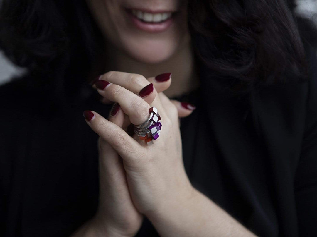 Een glimlachend persoon met donker haar vouwt zijn handen, accentueert kastanjebruine nagellak en draagt de Ring Kubusjes Groen R4GR van CLIC Creations. De wazige achtergrond accentueert het unieke geometrische ontwerp van de ring.