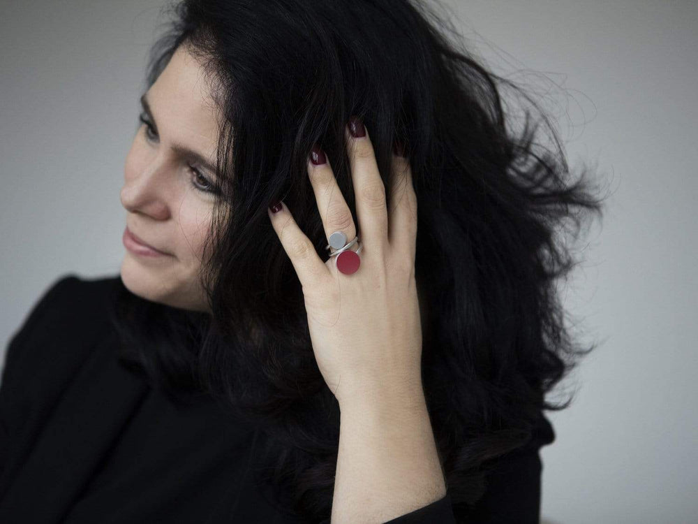 Een persoon met lang zwart haar raakt zijn hoofd aan en draagt een CLIC Creations ring, Twee ronde vormen Roze R5RO. Dit handgemaakte in Nederland stuk heeft een uniek ontwerp met twee ronde vormen in zilver en roze. Ze complementeren deze opvallende look met een zwarte outfit en donkere nagellak tegen een grijze achtergrond.
