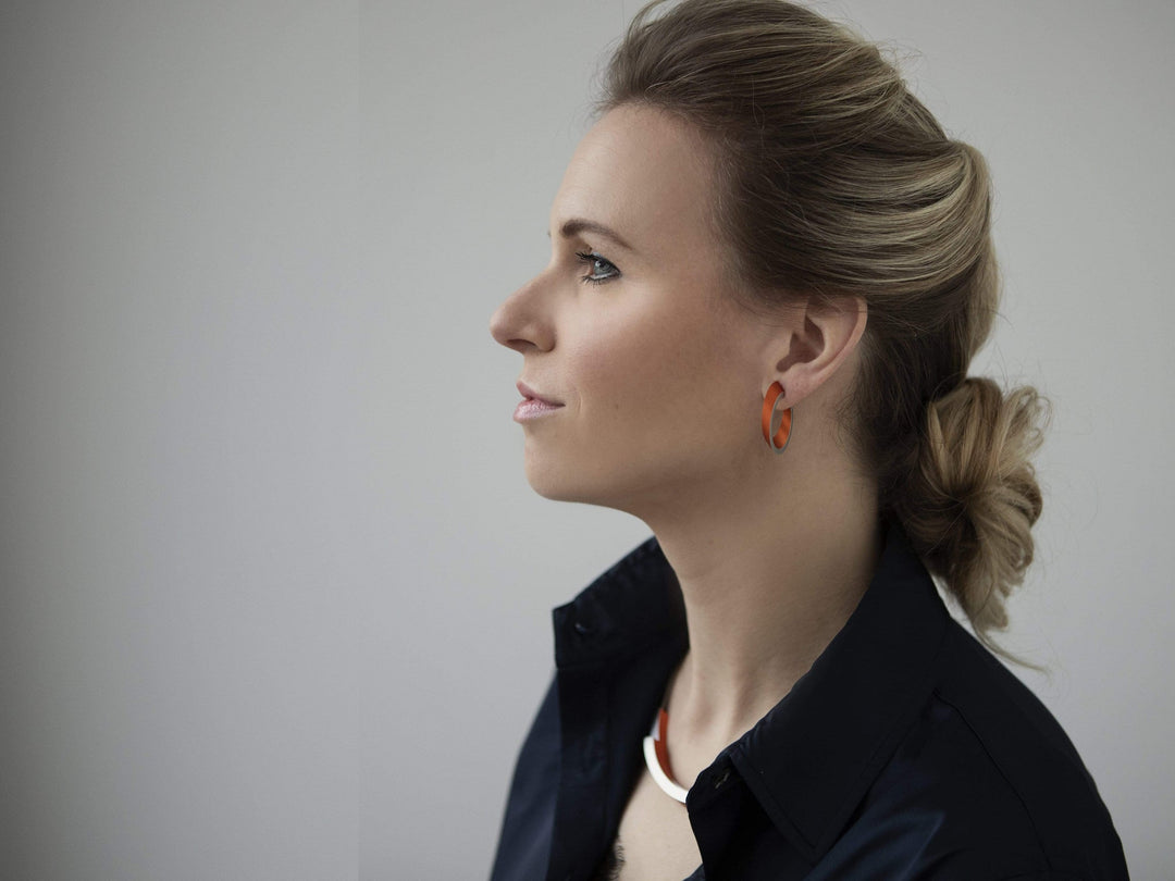 Profiel van een vrouw met licht haar in een elegante knot, oranje oorbellen en een donkere blouse met open kraag, die haar CLIC Creations Collier Bogen Goud|Geel C74G laat zien. Ze staat tegen een neutrale achtergrond en straalt evenwicht en verfijning uit.