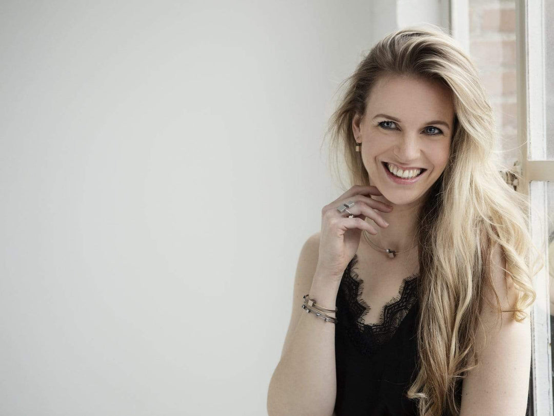 Een vrouw met lang blond haar lacht, gekleed in Clic creations A231G zwarte kanten top, staande bij een raam. Haar hand, versierd met ringen en armbanden, rust bij haar kin terwijl ze recht in de camera kijkt tegen een lichte, minimalistische achtergrond.