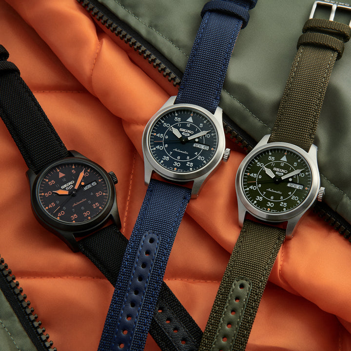 Op een gewatteerd oppervlak met een oranje en groene ritsachtergrond worden drie polshorloges met stoffen bandjes tentoongesteld: een zwart horloge met oranje accenten, een blauw horloge met zilveren kast en een groen ontwerp in militaire stijl. De Seiko 5 Sports SRPH29K1 benadrukt het befaamde vakmanschap van het merk.