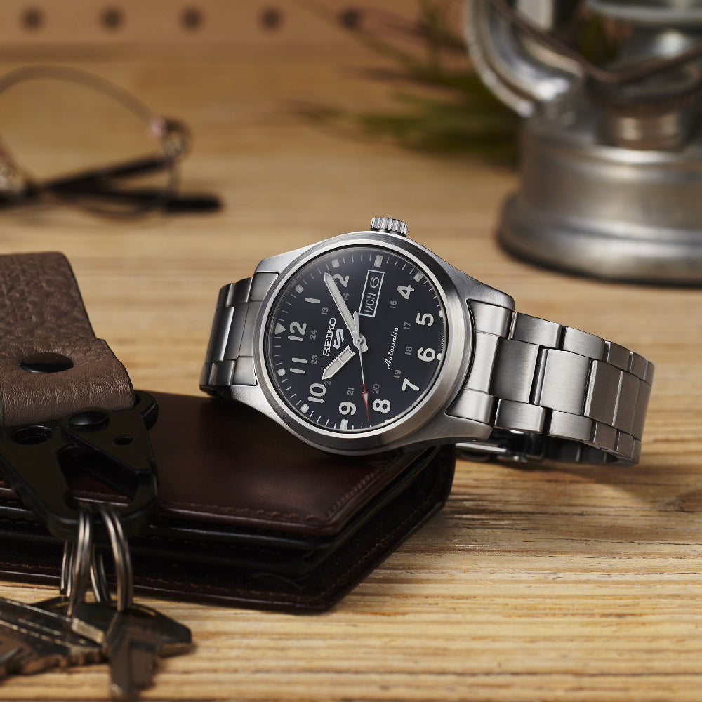 Een Seiko 5 Sports herenhorloge SRPG27K1 met een zilveren metalen band en zwarte wijzerplaat rust op hout, naast een leren portemonnee en een set sleutels, onder het zachte licht van een lamp. Perfect stijlvol.