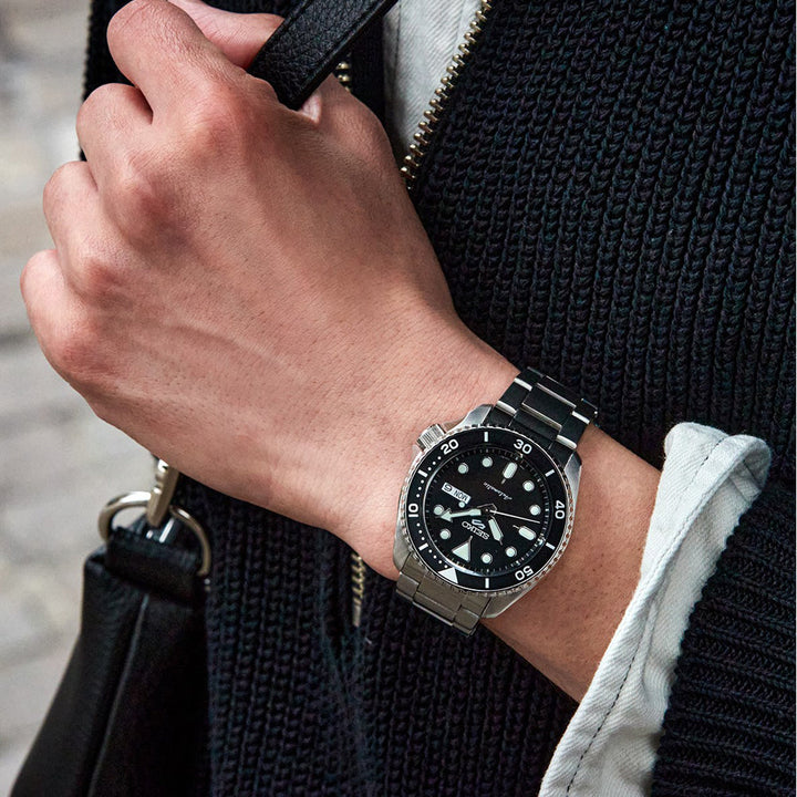 Een close-up van iemand die het Seiko 5 Sports heren horloge SRPD55K1 by Seiko draagt, toont de zwarte wijzerplaat, witte markeringen en roestvrijstalen kast op 10:10. De persoon houdt een zwarte tas vast terwijl hij een gebreide zwarte trui over een wit shirt draagt.