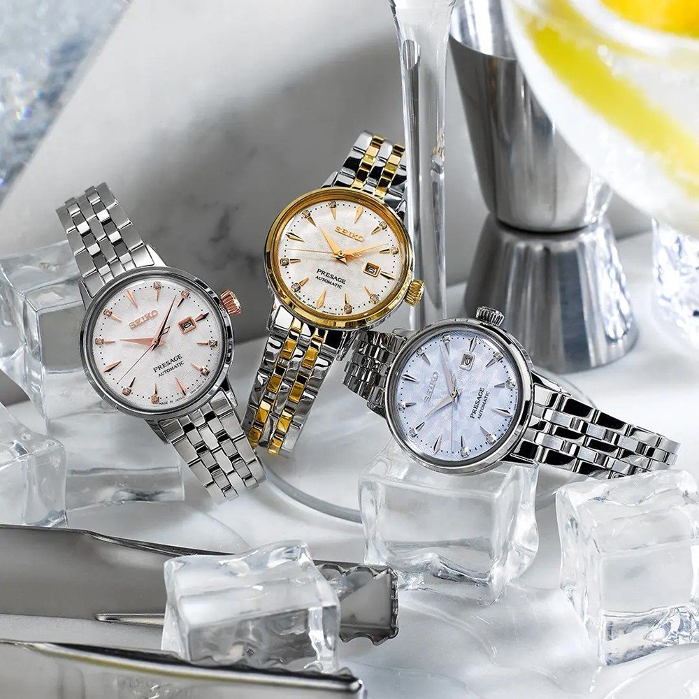 Drie elegante polshorloges met metalen banden rusten op ijzige kubussen, elk met unieke bezels: zilver, goud en goud-geaccentueerd. Het Seiko Presage dameshorloge SRE009J1 voegt verfijning toe met zijn Hardlex mineraalglass voorkant. Een cocktailshaker en een schijfje citroen verschijnen op de achtergrond.