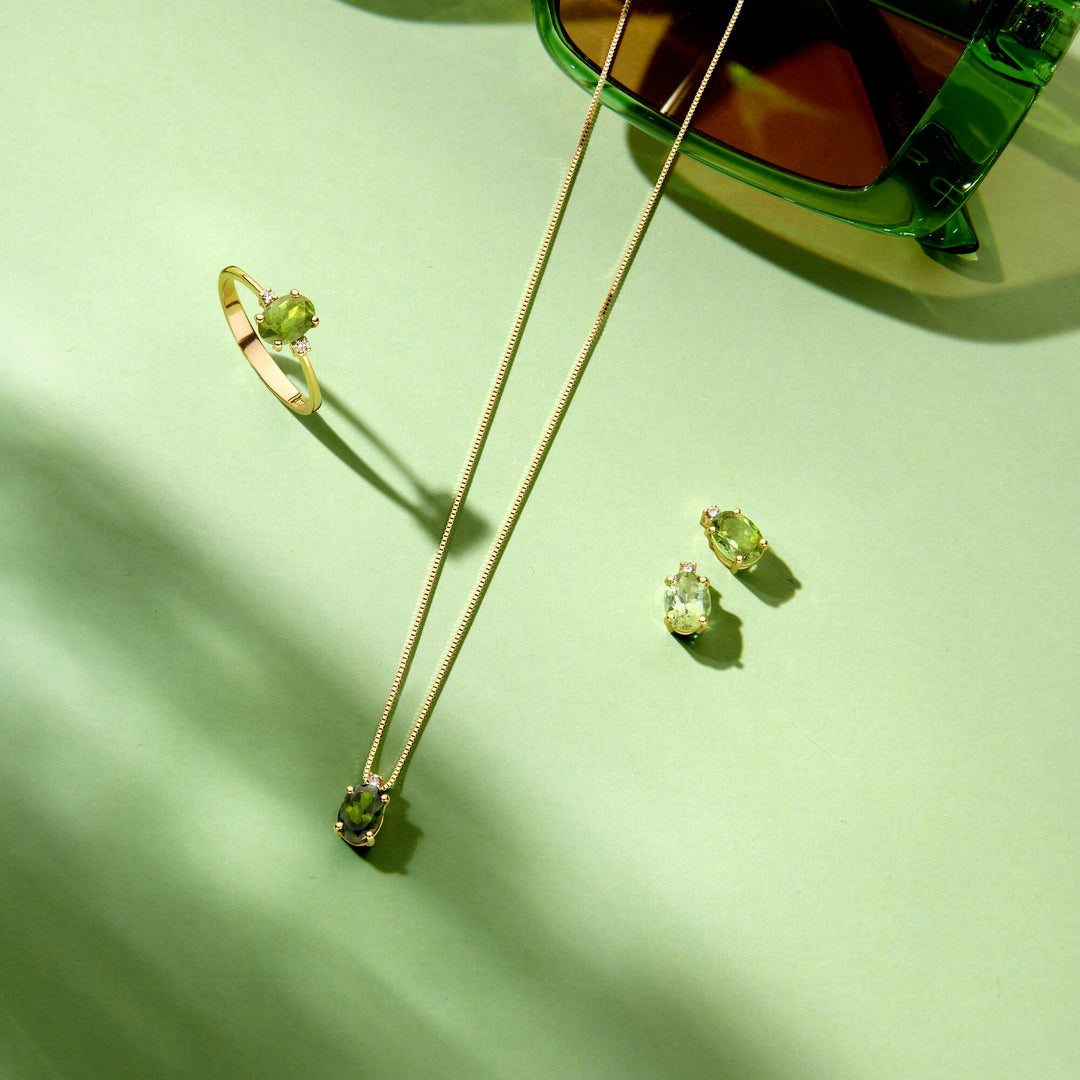 Een Van der Kooij Collectie 14K geelgouden trilogie-ring met toermalijn en diamant van 0.04ct H SI ligt op een lichtgroene ondergrond naast groenomrande zonnebrillen.