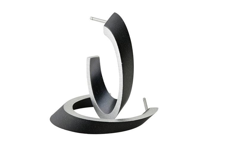 De Ovale creolen Zwart O21Z van CLIC Creations zijn zwarte, gebogen oorbellen met zilveren randen, gemaakt van hypoallergeen chirurgisch staal. Ze hebben een strak, modern design en zijn gepositioneerd alsof ze tegen elkaar aan leunen.