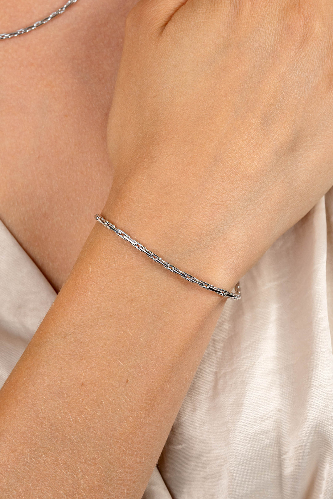 Een close-up toont de pols van een persoon die de Zinzi Armband zilver ZIA2648 (18-21cm) draagt. De elegante, zilveren gedraaide armband van Zinzi contrasteert prachtig met hun lichte, satijnachtige kleding terwijl hun hand vlakbij het sleutelbeen rust.