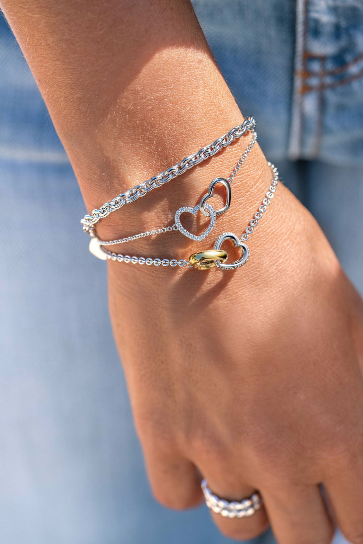 Een hand versierd met drie zilveren Zinzi-armbanden met hartontwerpen en een Zinzi Armband Zilver Zia1911, gecombineerd met een glimmende zilveren ring, past perfect bij een spijkerbroek, omdat de sieraden schitterend glinsteren in het licht.