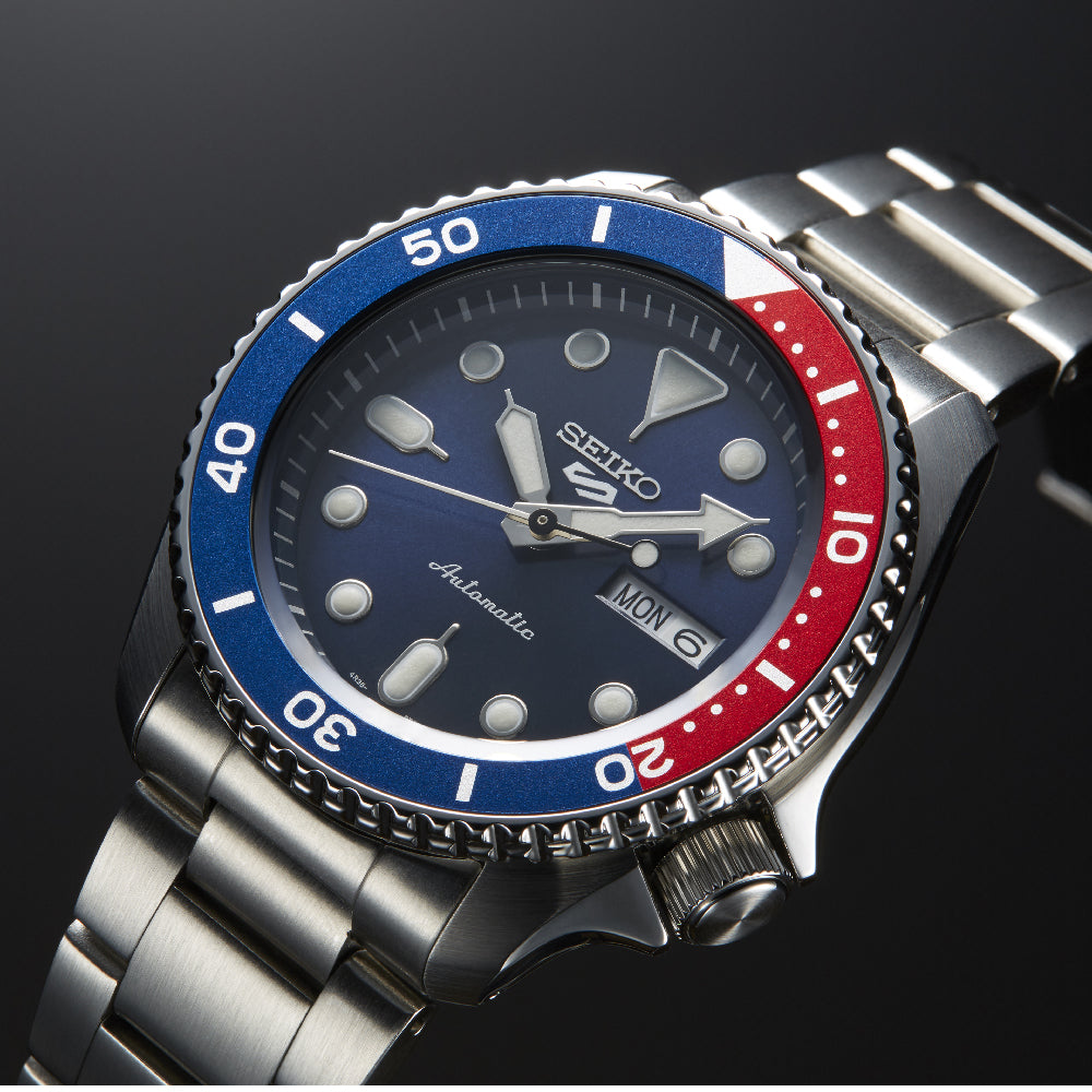 Een close-up van de Seiko 5 Sports heren horloge SRPD53K1 van Seiko, met een metalen band, blauwe en rode lunette, donkerblauwe wijzerplaat met lichtgevende markeringen en een automatisch uurwerk met dag-datum aanduiding (MON/6) op een zwarte achtergrond.